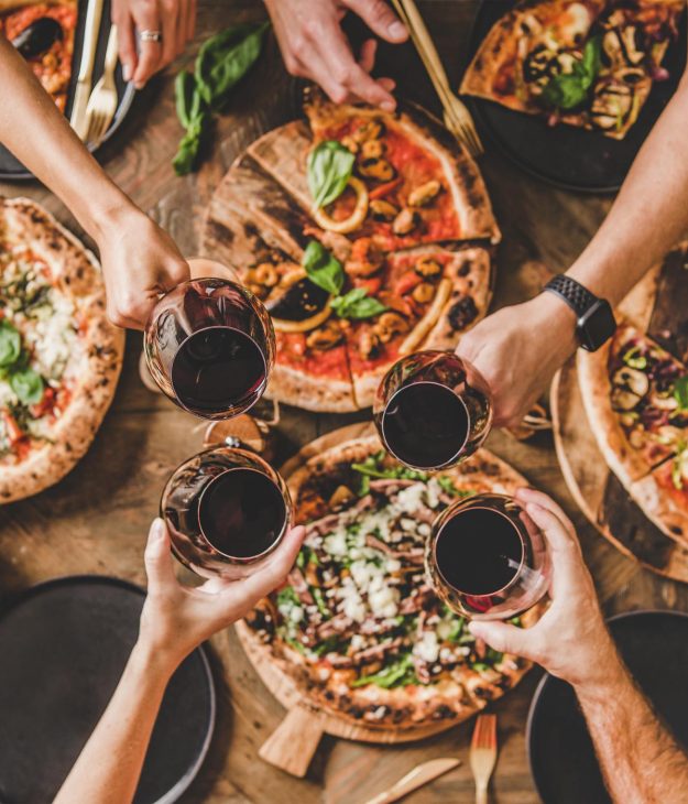 Family or friends having pizza party dinner. Flat-lay of people clinking glasses with red wine over rustic wooden table with various kinds of Italian pizza, top view. Fast food lunch, celebration
